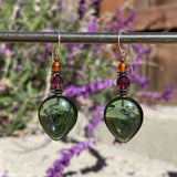 Green teardrop earrings with brown and purple beads on a metal rod against a blurred natural background.