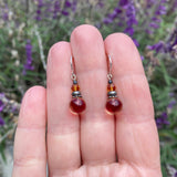 Pair of red beaded earrings held between fingers with a blurred purple flower background