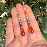 A pair of colorful beaded dangle earrings displayed on a person's hand, with green leaves in the background. The earrings feature silver french hooks, small faceted beads in orange, light blue, yellow-green, and magenta, and a large teardrop-shaped orange drops at the bottom.