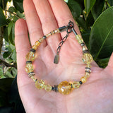 A beaded bracelet with faceted yellow beads and dark metal accents, displayed on an open hand with green leaves in the background.