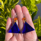 Blue triangle earrings with orange beads held in a hand against a green leafy background