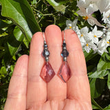 Purple  beaded earrings held between fingers with a natural background of leaves and flowers