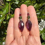 Amethyst earrings with silver and red accents held in a hand against a green leafy background