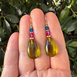 A pair of handmade teardrop dangle earrings held in a hand. The earrings feature gold-filled metal ear wires, small blue and pink faceted beads, and a large polished olive-yellow teardrop bead at the bottom.