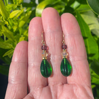 Green teardrop earrings with gold and red accents held between fingers against a green leafy background
