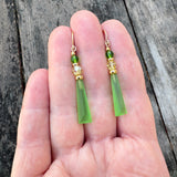Green earrings with gold accents held between fingers against a wooden background