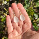 A pair of teardrop-shaped white glass drop earrings with gold hooks and clear faceted beads, held in a person's hand against a blurred green outdoor background.