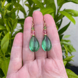 Green teardrop earrings with pink beads held in a hand against a green leafy background