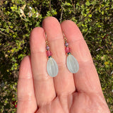 A pair of handmade dangle earrings held in a person's hand, featuring pale greenish-white teardrop-shaped beads, pink textured spacer beads, and small gold-toned metal details, with a background of green foliage.