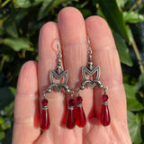 Silver earrings with red teardrop beads held in a hand against a green leafy background