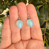 A pair of teardrop-shaped iridescent light blue glass beads on silver fishhook wires, held in a person's hand with green leaves in the background.