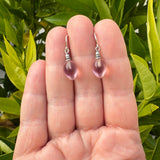 A pair of silver drop earrings with teardrop-shaped light pink gemstones, held on a person's fingers with green leaves in the background.