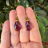 A pair of teardrop-shaped dangling earrings with faceted burgundy-purple stones and small gold-toned beads, held between a person's fingers with green leaves in the background.