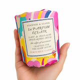 A hand holding a colorful Stannie & Lloyd La Parfum Éclair perfume candle box against a plain white background.