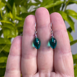 Teal drop earrings held between fingers with a green leafy background