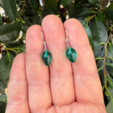 A pair of green teardrop-shaped glass drop earrings with silver fishhook backs, held on a person's fingers against a background of green leaves.