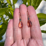 Pair of peach-colored teardrop earrings on a hand with a green leafy background