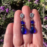 Blue teardrop earrings with silver tops held between fingers against a blurred natural background