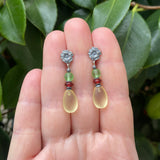 Pair of earrings with floral tops and beaded elements held between fingers against a green leafy background
