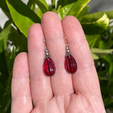 Red teardrop earrings held between fingers with a green leafy background
