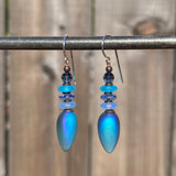 Blue teardrop earrings with beaded accents hanging on a metal rod against a blurred natural background.