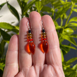 Orange teardrop earrings held between fingers with a green leafy background