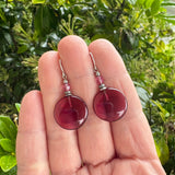 Red earrings held in a hand with a green leafy background