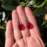 Red drop earrings held between fingers against a leafy background