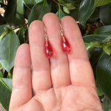 A pair of gold-tone drop earrings with translucent pinkish-red teardrop-shaped beads, shown resting on an open hand with green leaves in the background.