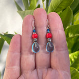 Pair of earrings with red and clear beads held between fingers against a green leafy background