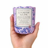 A hand holding a Stannie & Lloyd La Parfum & Clair scented candle in a lavender container with a blue floral pattern and a white label.