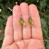 A pair of small teardrop-shaped glass earrings held on a person's fingers with green leaves in the background.