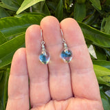 A pair of dangling earrings with teardrop-shaped blue and pink glass beads and silver hooks, held between a person's fingers with green leaves in the background.