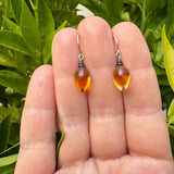 A pair of teardrop-shaped topaz glass drop earrings with silver hooks, held between a person's fingers against a background of green leaves.