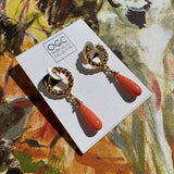 A pair of gold huggie hoop earrings with coral orange teardrop drops, displayed on a white card set against a colorful abstract background.