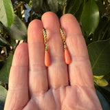 A pair of gold and coral orange drop earrings held on a person's fingers with green leaves in the background.
