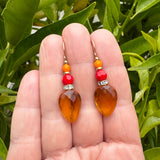 A pair of dangling earrings held in a person's hand, featuring faceted amber-orange teardrop beads, red beads, and small gold-toned metal details, with green leaves in the background.