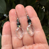 A pair of dangling earrings with faceted clear teardrop-shaped beads, black faceted beads, and small iridescent accent beads, shown resting on an open hand with dark green leaves in the background.