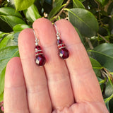 Red beaded earrings held between fingers with a green leafy background