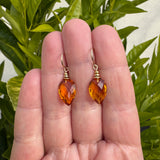 Topaz faceted glass earrings with gold trim, held between fingers in front of a lime tree.