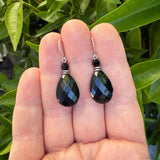 A pair of faceted black teardrop dangle earrings with silver hooks, held between a person's fingers with green leaves in the background.