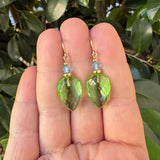 A pair of gold drop earrings with faceted light green teardrop-shaped gemstones and small round blue beads, held between a person's fingers with green leaves in the background.