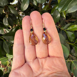 A pair of gold drop earrings with faceted brown teardrop gemstones and small black beads, held in a person's hand with green leaves in the background.