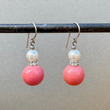 A pair of dangling earrings with silver wires, a small white pearl, a silver spacer with clear stones, and a large round pink bead drop below, hanging from a black rod against a plain gray background.