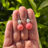A pair of dangling earrings held between fingers outdoors, with green foliage blurred in the background. Each earring has a silver earwire, a small white pearl bead, and a round coral-pink bead below.