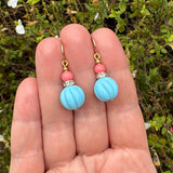 A pair of handmade dangle earrings held in a person's hand, featuring light blue ribbed beads, pink coral beads, and gold hooks against a background of green foliage.