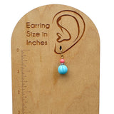 A wooden earring size display card with a laser-engraved ear profile, inch ruler, and text reading 'Earring Size in Inches.' A single dangling earring is shown hanging from the ear-shaped notch: a small pink bead, a gold spacer ring, and a round light blue bead.