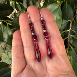 A pair of dangling earrings with faceted dark red beads and silver-toned spacers, held in a person's hand against a background of green leaves.