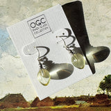Pair of yellow glass earrings on a card with 'OGC Owen Glass Collection' branding, set against a scenic background.