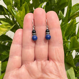 Blue and black Czech glass earrings with jet rhinestones. Earrings are held in a hand with greenery in the background. 